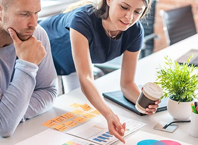 Modern ofis ortamında çalışan iki kadın, masa üzerinde karton bardak örnekleri ve tasarım çizimleriyle birlikte ürün geliştirme sürecini planlıyor. Yaratıcı ekip çalışmasına odaklanılmış.